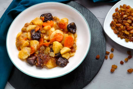 A dish of Jewish cuisine sweet tsimes with carrots vegetarian dates in a plate on a round stand on a concrete background next to raisins on a saucer.の写真素材