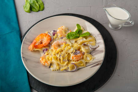 Farfalle pasta with shrimps in a creamy sauce on a gray plate on a concrete background next to a blue napkin and a jug of cream. Horizontal photoの写真素材