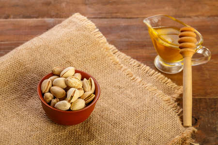 Pistachios in a clay bowl on a sacking next to honey with a spoon on a wooden table Horizontal photoの写真素材