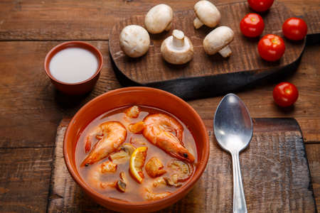 Tom yam soup with shrimps in a plate on a wooden table with a spoon next to a bowl with coconut milk mushrooms and tomatoes.の写真素材