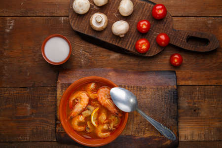 Tom yam soup with shrimps in a plate on a wooden table with a spoon next to a bowl with coconut milk mushrooms and tomatoes.の写真素材