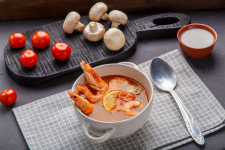Tom yam soup with shrimps in a tureen with a spoon on a concrete background next to a bowl with coconut milk mushrooms and tomatoes.の写真素材