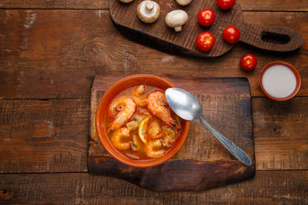 Tom yam soup with shrimps in a plate on a wooden table on a stand with a spoon next to a bowl of coconut milk mushrooms and tomatoes.の写真素材
