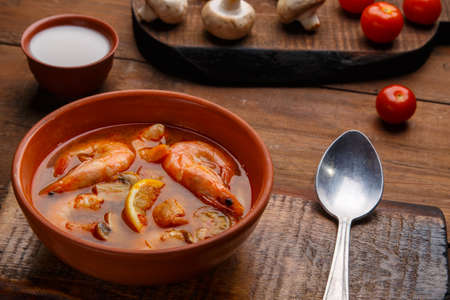Tom yam soup with shrimps in a plate on a wooden table on a stand with a spoon next to a bowl of coconut milk mushrooms and tomatoes.の写真素材