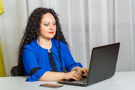 Curly brunette woman sitting at a table in the office working with a laptop.の写真素材