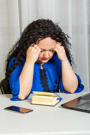 Curly brunette woman sits at the table in the office with her head on her hands.の写真素材