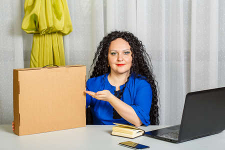 Curly joyful brunette woman sits at a table in the office holding a copy of a place purchase in her...