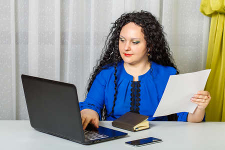 Curly brunette woman sits at the table in the office prints the text from the sheet.の写真素材