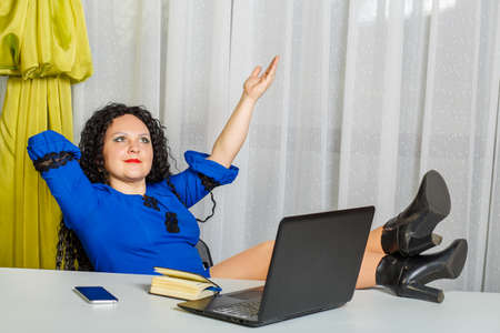 Curly brunette woman sits at a table in the office resting after finishing work with her legs on the table.の写真素材