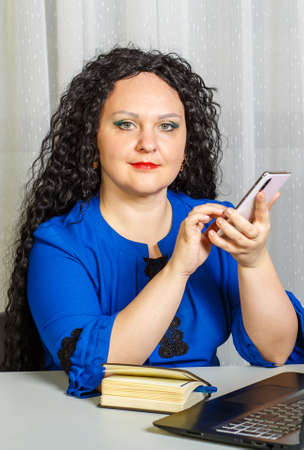 Curly brunette woman sits at a table in the office working with a smartphone.の写真素材
