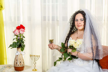 A happy Jewish bride sits in the hall before the chuppah ceremony at a table with flowers with a glass of wine for Kiddush and a bouquet of white roses.の写真素材