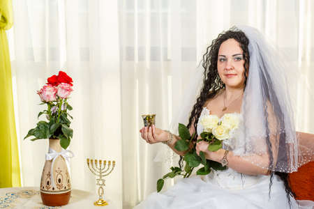 A happy Jewish bride sits in the hall before the chuppah ceremony at a table with flowers with a glass of wine for Kiddush and a bouquet of white roses.の素材 [FY310167230534]