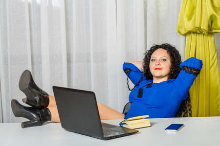 A curly-haired brunette in blue sits on a chair and puts her legs on the tableの写真素材