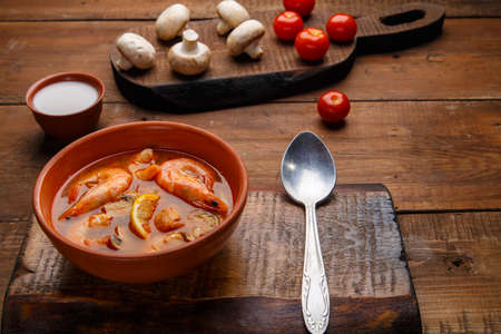 Tom yam soup with shrimps in a plate on a wooden table on a stand with a spoon next to a bowl of coconut milk mushrooms and tomatoes.の写真素材