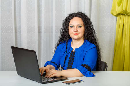 Curly brunette woman sitting at a table in the office working with a laptop.の写真素材