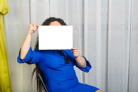 Curly joyful brunette woman on a chair holds a copy space plate in her hands, covering her face.の写真素材