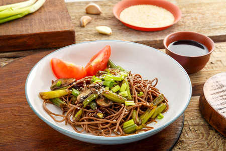 Soba with mushrooms, green beans and sesame seeds in a plate on a stand on a wooden table near chopsticks and ingredients.の写真素材
