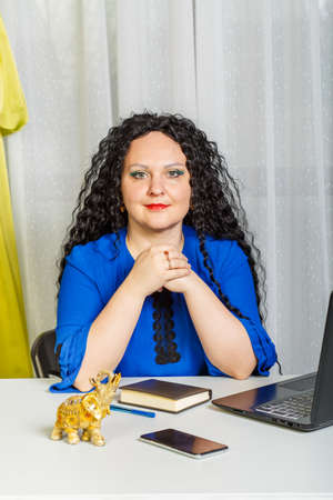 Curly brunette woman sits at the table in the office with a laptop.の写真素材