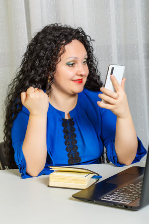Curly brunette woman sits at a table in the office working with a smartphone.の写真素材