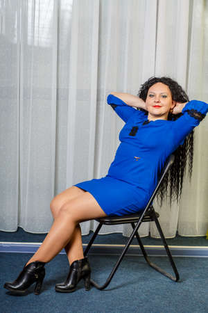 Curly brunette woman in blue sits on a chair and points to something with her hand.の写真素材
