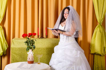 A Jewish bride in a white wedding dress with a veil stands in the hall at a table with flowers and prays before the chuppah ceremony. Horizontal photoの写真素材
