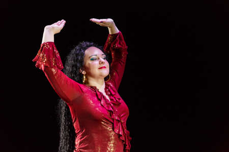 A gypsy woman with curly black hair dances in a red dress on a black background. Horizontal photoの写真素材