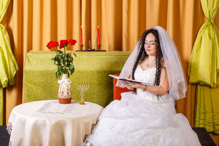 A Jewish bride in a white wedding dress with a veil sits at a table with flowers and reads blessings from a prayer book before the chuppah ceremony. Horizontal photoの写真素材