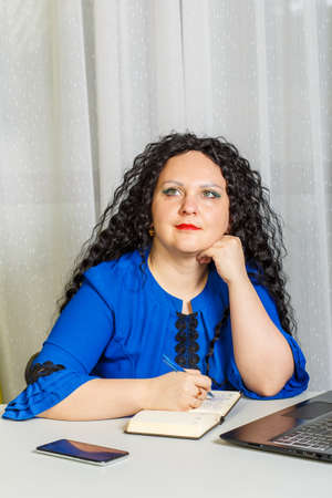 Curly pensive brunette woman is sitting at the table in the office. Vertical photoの写真素材