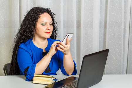 Curly brunette woman sits at the table in the office talking on the phone.の写真素材