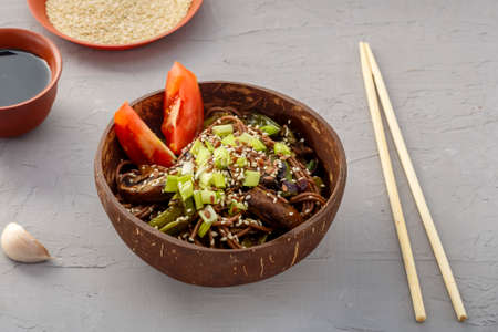 Soba with mushrooms and sesame seeds in a plate of coconut shells on a concrete background near garlic and soy sauce and sticks. Horizontal photoの写真素材