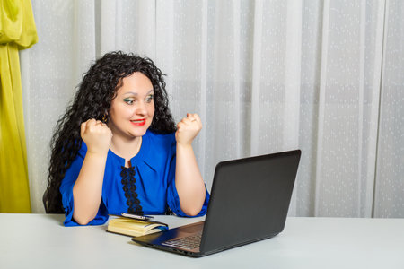 Curly brunette woman sits at a table in the office emotionally communicates via video communication. Horizontal photoの写真素材