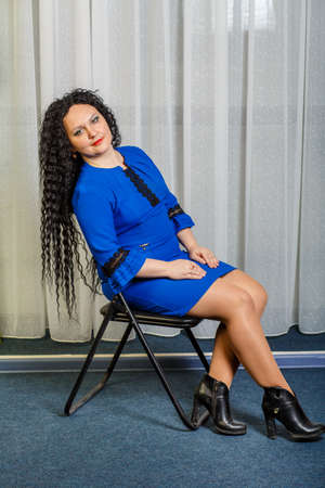 Curly brunette woman in blue sits on a chair with her head thrown back and stretched her legs. Vertical photoの写真素材