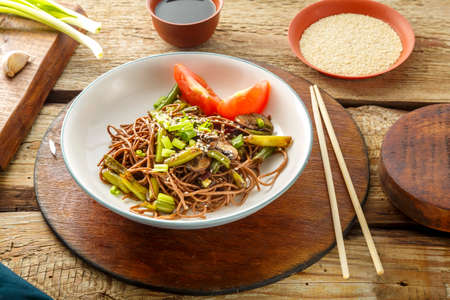 Soba with mushrooms, green beans and sesame seeds in a plate on a stand on a wooden table next to chopsticks and soy sauce and garlic. Horizontal photoの写真素材