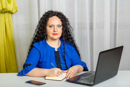 Curly brunette woman sitting at a table in the office working with a laptop.の写真素材