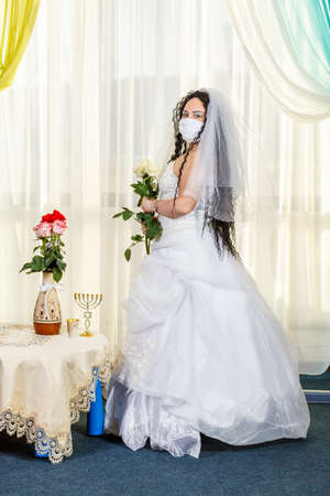 A Jewish bride sits waist-deep in a synagogue at a table with flowers before a chuppa ceremony during a pandemic, wearing a medical mask and a bouquet of flowers, awaits the groom.の写真素材