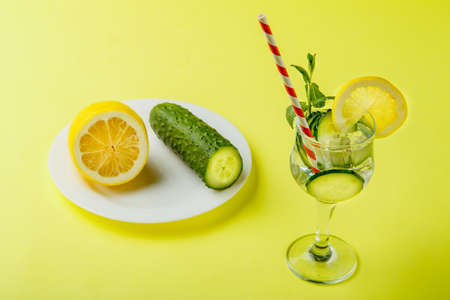 Cocktail cucumber water with lemon and mint in a glass on a napkin on a yellow background next to cucumbers and lemon on a napkin. Horizontal photoの写真素材