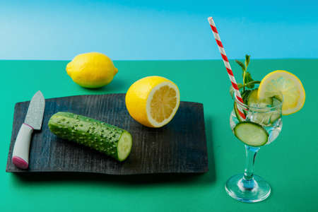 Cocktail cucumber water with lemon and mint in a glass on a napkin on a green background next to a lemon and cucumbers on a napkin and a knife. Horizontal photoの写真素材