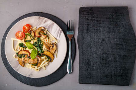 Udon with mushrooms and spinach in a plate on a concrete background near a fork and a wooden board. Horizontal photoの写真素材