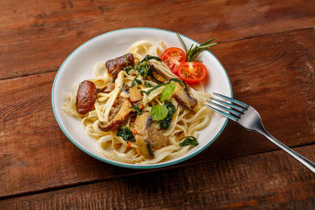 Udon with mushrooms and spinach and cherry tomatoes in a plate on a wooden table next to a fork. Horizontal photoの写真素材