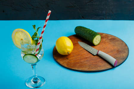 Cocktail cucumber water with lemon and mint in a glass on a napkin on a blue background next to cucumbers and a knife. Horizontal photoの写真素材