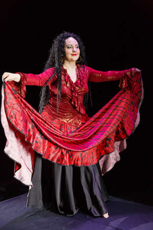 Gypsy woman barefoot with long black hair dances waving red-black skirt on a black background. Vertical photoの写真素材