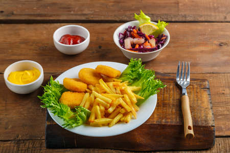 French fries with chicken nuggets next to cheese sauce and ketchup in a gravy boat and salad on a wooden table next to a fork. Horizontal photoの写真素材