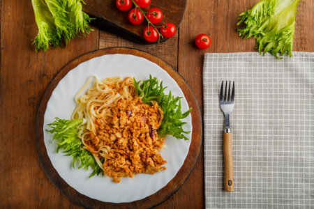 A plate of bolognese pasta decorated with greens on a wooden table next to a gray napkin and a fork and cherry tomatoes. Horizontal photoの写真素材