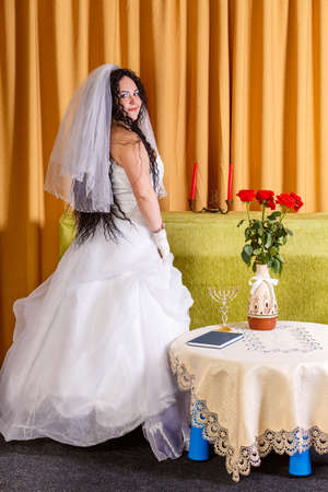 A Jewish bride in a white wedding dress with a veil stands in the hall at a table with flowers half-turned before the chuppah ceremony. Vertical photoの写真素材