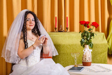 A Jewish bride in a white wedding dress with a veil sits at a table with flowers communicating in instant messengers before the chuppah ceremony. Horizontal photoの写真素材