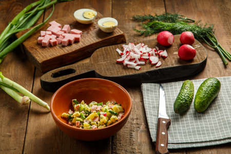Okroshka in a clay plate on a table on a wooden table next to an egg sausage and cucumbers on planks. Horizontal photoの写真素材