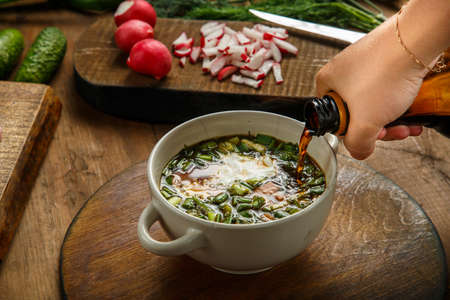 A woman's hand pours kvass from a bottle into okroshka in a tureen with herbs and an egg. Horizontal photoの写真素材