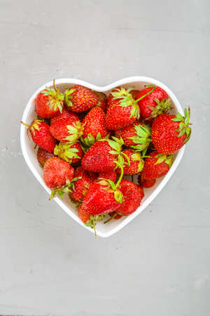 Ripe strawberries in a plate in the shape of a heart on a gray background. Vertical photoの写真素材