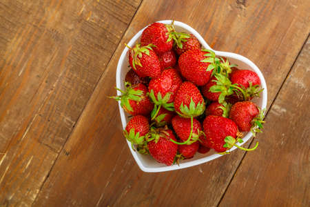 Fresh large strawberries in a white plate in the shape of a heart on the table. Horizontal photoの写真素材