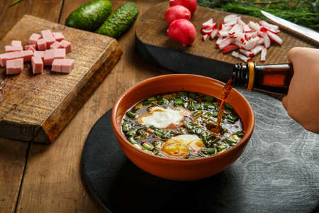 A female hand pours kvass from a bottle into okroshka with herbs and an egg. Horizontal photoの写真素材